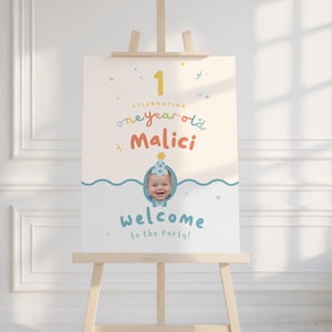 Puede incluir: Un letrero de bienvenida para una fiesta de primer cumplea&ntilde;os, exhibido en un caballete de madera. El letrero presenta el texto "1", "celebrating one year old Malici" y "welcome to the party!" en fuentes coloridas y divertidas. Se incluye una foto de un beb&eacute; con un gorro de fiesta.