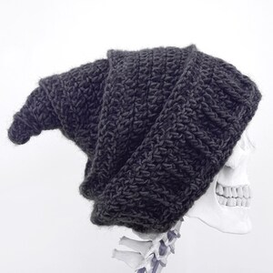 May include: A dark grey, chunky knit beanie hat. The hat is draped over a white human skull, with the teeth and part of the spine visible. The background is plain white.