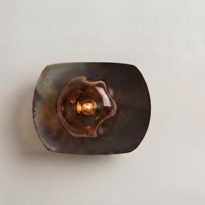 May include: A wall-mounted light fitting featuring a rectangular, dark, iridescent metal backplate with a slightly curved, organic shape. Centred on the plate is a translucent, amber-coloured glass shade with a wavy, flower-like form.