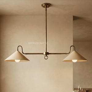 Puede incluir: Una lámpara colgante de dos luces en tono latón. La lámpara tiene una varilla vertical central con una barra horizontal que se extiende a cada lado. Cada lado tiene una pantalla cónica de color crema que apunta hacia abajo con una bombilla.