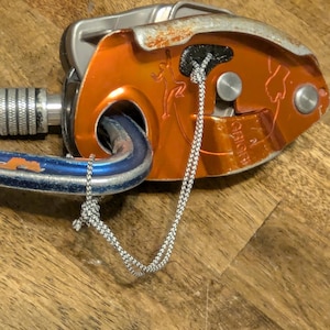 May include: An orange climbing belay device with a silver top and a blue and silver carabiner. A white and gray rope is attached to the device. The device has a climber graphic.