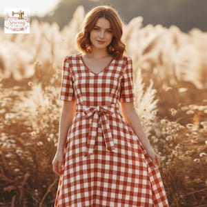 May include: A red and white checkered dress with a V-neck and short sleeves. The dress has a matching tie belt at the waist. The dress is knee-length and is worn by a person standing in a field of tall grass.