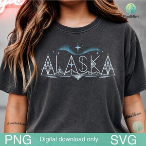 Puede incluir: Una camiseta gris carbón de manga corta con un diseño gráfico en blanco y azul claro. El estampado muestra la palabra ALASKA en una fuente geométrica, acentuada con picos de montaña estilizados, un reflejo, estrellas y un patrón de aurora boreal.