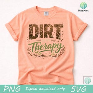 Puede incluir: Camiseta color melocot&oacute;n con mangas enrolladas. La camiseta presenta el texto "DIRT is my Therapy" en un dise&ntilde;o texturizado y desgastado. La palabra "Therapy" est&aacute; en verde con un borde de hojas e ilustraciones de peque&ntilde;as plantas.