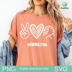 May include: A coral-coloured t-shirt featuring a white graphic design of a peace sign, a heart, and two hands clasped together. Below the graphic, the text #ENDBULLYING is printed in white. The shirt is worn with a blue denim jacket.
