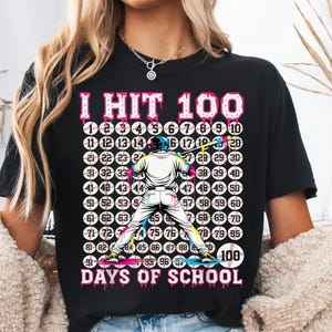 Peut inclure: T-shirt noir avec l'inscription "I HIT 100 DAYS OF SCHOOL" en rose et blanc. Le motif comprend un joueur de baseball et une grille de balles de baseball numérotées de 1 à 100. C'est un vêtement décontracté.