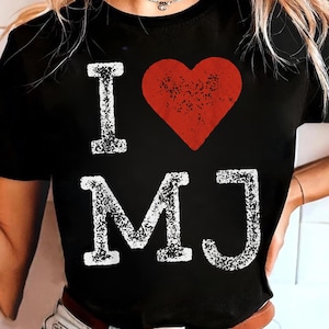 Peut inclure: T-shirt noir avec l'inscription "I ♥ MJ" en blanc, un cœur rouge remplaçant le mot "love". Les lettres ont un aspect texturé et vieilli. Le t-shirt est de style col rond.