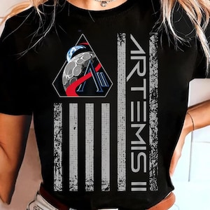 Può includere: T-shirt nera con un design grafico relativo alla missione spaziale Artemis II. Il design include un emblema triangolare con una luna e la Terra, il logo Artemis II e strisce verticali che assomigliano a una bandiera.