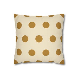 Pode incluir: Almofada decorativa quadrada com fundo creme e grandes bolinhas marrom dourado espaçadas uniformemente. A almofada tem uma aparência macia e texturizada, sugerindo um item de decoração confortável.