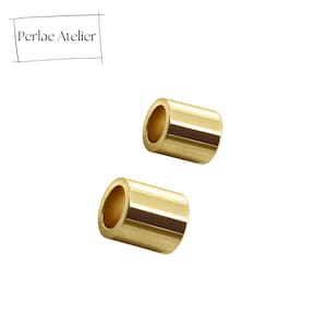 May include: Two gold-colored, cylindrical beads with a polished finish. The beads have a hollow center and are displayed against a white background. The upper left corner of the image features the text "Perlae Atelier" in a black font.