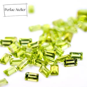 Peut inclure: Plusieurs petites pierres précieuses rectangulaires vert clair sont dispersées sur une surface blanche. Les pierres précieuses scintillent légèrement et sont orientées différemment. Le texte "Perlae Atelier" est dans un encadré noir en haut à gauche.