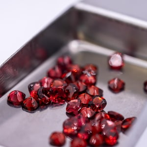 Pode incluir: Uma coleção de pedras preciosas facetadas, vermelho-escuras, é exibida numa pequena pá de metal prateado. As gemas, com corte brilhante, refletem a luz. A pá está sobre um fundo branco, realçando a cor e a clareza das gemas.