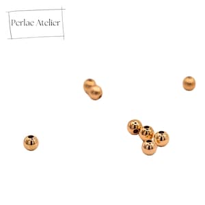 Könnte beinhalten: Mehrere kleine, runde, goldfarbene Perlen auf weißem Hintergrund. Die Perlen haben einen metallischen Glanz und scheinen aus Metall zu sein. Der Text "Perlae Atelier" befindet sich in der oberen linken Ecke.