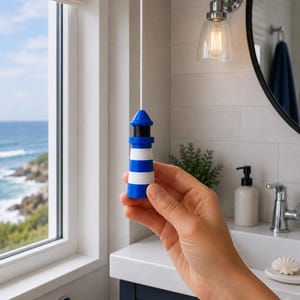 May include: A lighthouse-shaped light pull cord handle featuring horizontal blue and white stripes, a black middle section, and a blue conical top, attached to a white cord for operating a light switch in a bathroom setting.