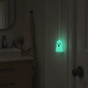 May include: A glowing, light-green ghost-shaped light pull attached to a white cord. The ghost figure features two small circular eyes and an oval-shaped mouth, emitting a soft, luminous light against a dark, neutral-toned interior wall.