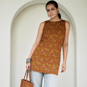 May include: A sleeveless tunic in a rust-brown color featuring a repeating yellow floral pattern, paired with light-wash blue denim jeans and a solid brown leather tote bag with two thin handles, worn with dark blue embroidered flat shoes.