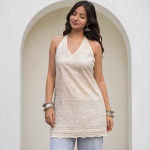 May include: A sleeveless cream-colored halter top featuring intricate white floral embroidery and a scalloped hemline, paired with light blue denim wide-leg trousers. The top has a V-neckline and detailed needlework patterns throughout the fabric.