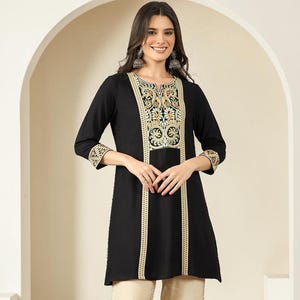 May include: A black tunic featuring three-quarter sleeves and a central panel with intricate gold, green, and white embroidery, paired with wide-leg beige trousers that have a subtle side trim detail and matching embellished open-toe sandals.