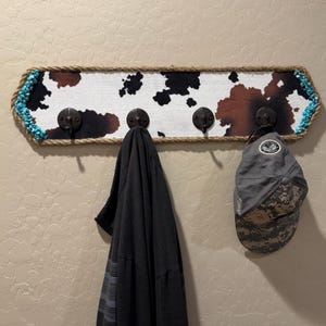 May include: A rectangular wall-mounted coat rack featuring a cowhide print pattern in black, white, and brown. The frame is wrapped in thick jute rope with turquoise stone accents at the corners. Four dark metal hooks hold a black shirt and a gray cap.