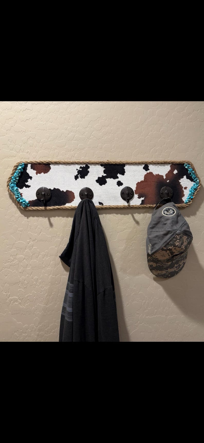 May include: A rectangular wall-mounted coat rack featuring a cowhide print pattern in black, white, and brown. The frame is wrapped in thick jute rope with turquoise stone accents at the corners. Four dark metal hooks hold a black shirt and a gray cap.