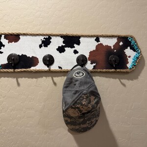 May include: A rectangular wall-mounted coat rack featuring a cowhide print pattern in black, white, and brown. The board is bordered by thick tan rope and turquoise accents, with four black metal hooks. A grey camouflage baseball cap hangs on one hook.