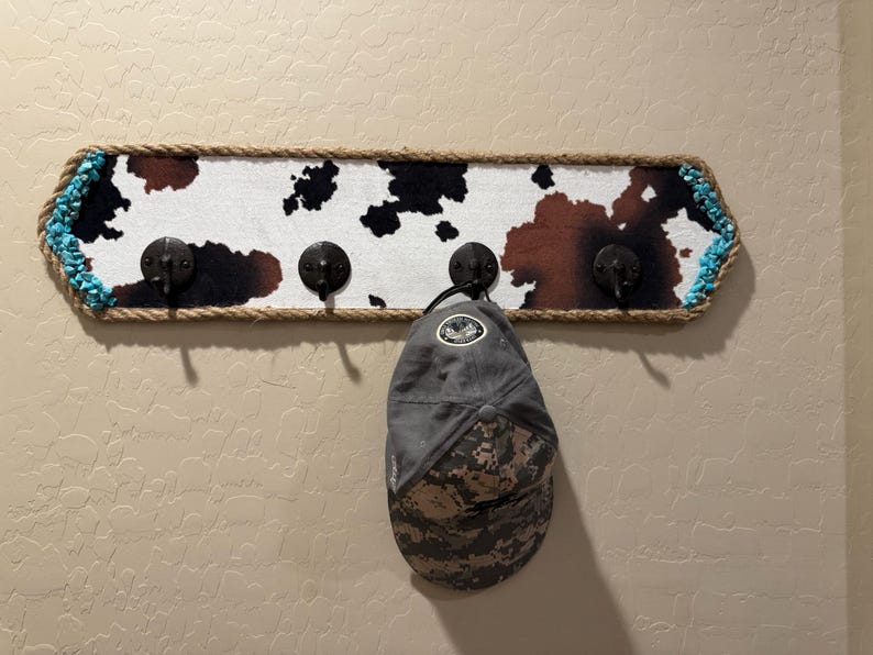 May include: A rectangular wall-mounted coat rack featuring a cowhide print pattern in black, white, and brown. The board is bordered by thick tan rope and turquoise accents, with four black metal hooks. A grey camouflage baseball cap hangs on one hook.