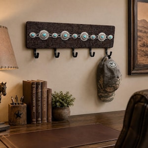 Puede incluir: Un perchero de pared rectangular con una base texturizada de color marrón oscuro adornada con una fila horizontal de adornos concho plateados, cada uno centrado con una pequeña piedra turquesa. El perchero incluye cinco ganchos de metal negro.
