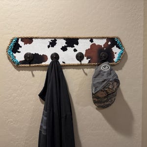 May include: A rectangular wall-mounted coat rack featuring a cowhide print pattern, bordered by thick tan rope and accented with turquoise beads at the ends. It includes four dark metal hooks, with a black garment and a gray baseball cap hanging.