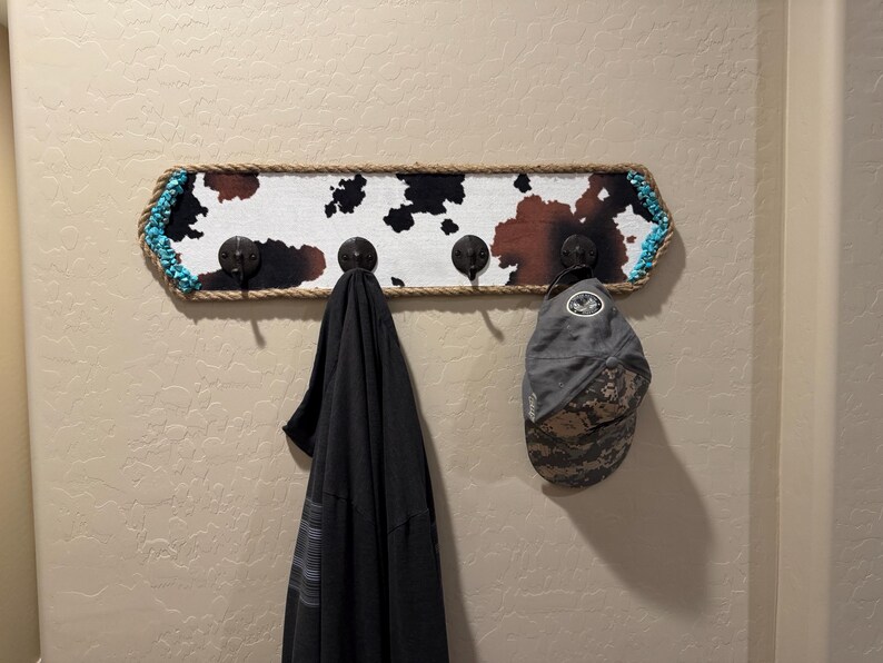 May include: A rectangular wall-mounted coat rack featuring a cowhide print pattern, bordered by thick tan rope and accented with turquoise beads at the ends. It includes four dark metal hooks, with a black garment and a gray baseball cap hanging.