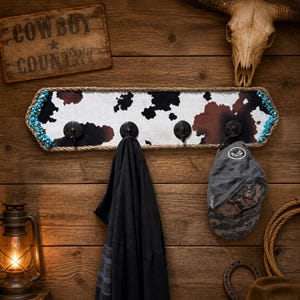 May include: A rectangular wall-mounted coat rack featuring a black and brown cowhide print pattern, bordered by thick rope and turquoise accents. The rack includes four black metal hooks, with a dark shirt and a grey baseball cap hanging from them.