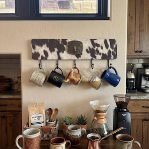 Puede incluir: Un soporte de pared con patrón de piel de vaca marrón y blanco y una hebilla central de metal con forma de longhorn. Cinco ganchos negros sostienen varias tazas de cerámica, incluyendo una azul y una con forma de cabeza de toro.