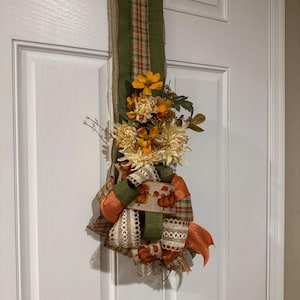 May include: A vertical door swag featuring a long strip of olive green and plaid burlap ribbon. The arrangement is decorated with artificial orange and cream-coloured flowers, green foliage, and a multi-layered bow with pumpkin-patterned ribbon.