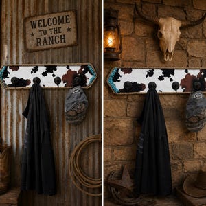 May include: A rectangular wall-mounted coat rack featuring a cowhide print pattern, bordered by a thick rope trim and turquoise accents. The rack includes four black metal hooks, with a black garment and a grey baseball cap hanging from them.