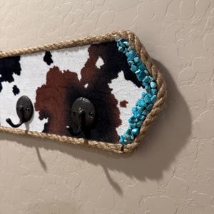 May include: A wall-mounted hook rack featuring a cowhide pattern in black, brown, and white. The rectangular board is bordered by thick, natural-colored rope and accented on the right side with small, bright turquoise stone chips.