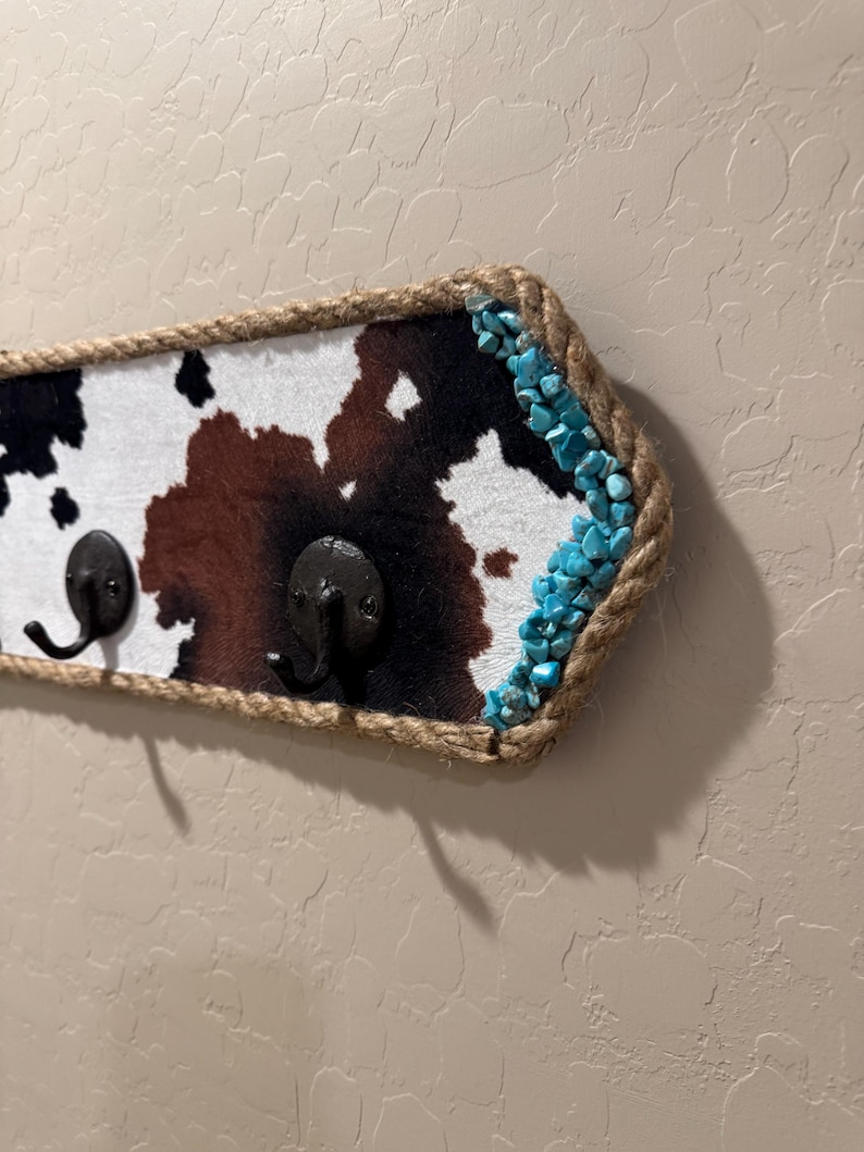May include: A wall-mounted hook rack featuring a cowhide pattern in black, brown, and white. The rectangular board is bordered by thick, natural-colored rope and accented on the right side with small, bright turquoise stone chips.