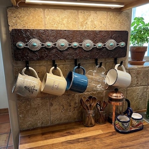 May include: A rectangular wall-mounted mug rack featuring a dark brown embossed leather-look panel decorated with a horizontal row of silver-toned conchos inlaid with small turquoise stones and five black metal hooks holding various mugs.