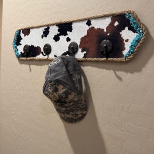 May include: A rectangular wall-mounted coat rack featuring a cowhide print pattern in brown, black, and white. The rack is bordered by thick jute rope and turquoise stones, with four dark metal hooks. A grey camouflage baseball cap hangs on one hook.