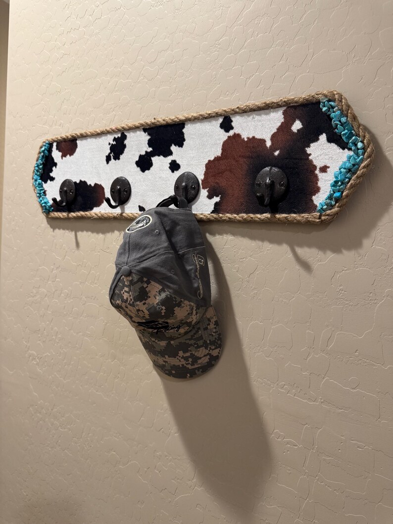 May include: A rectangular wall-mounted coat rack featuring a cowhide print pattern in brown, black, and white. The rack is bordered by thick jute rope and turquoise stones, with four dark metal hooks. A grey camouflage baseball cap hangs on one hook.