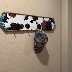 May include: A rectangular wall-mounted coat rack featuring a cowhide print pattern in black, brown, and white. The frame is wrapped in thick rope and accented with turquoise stones on the ends. Four black metal hooks hold a grey baseball cap.