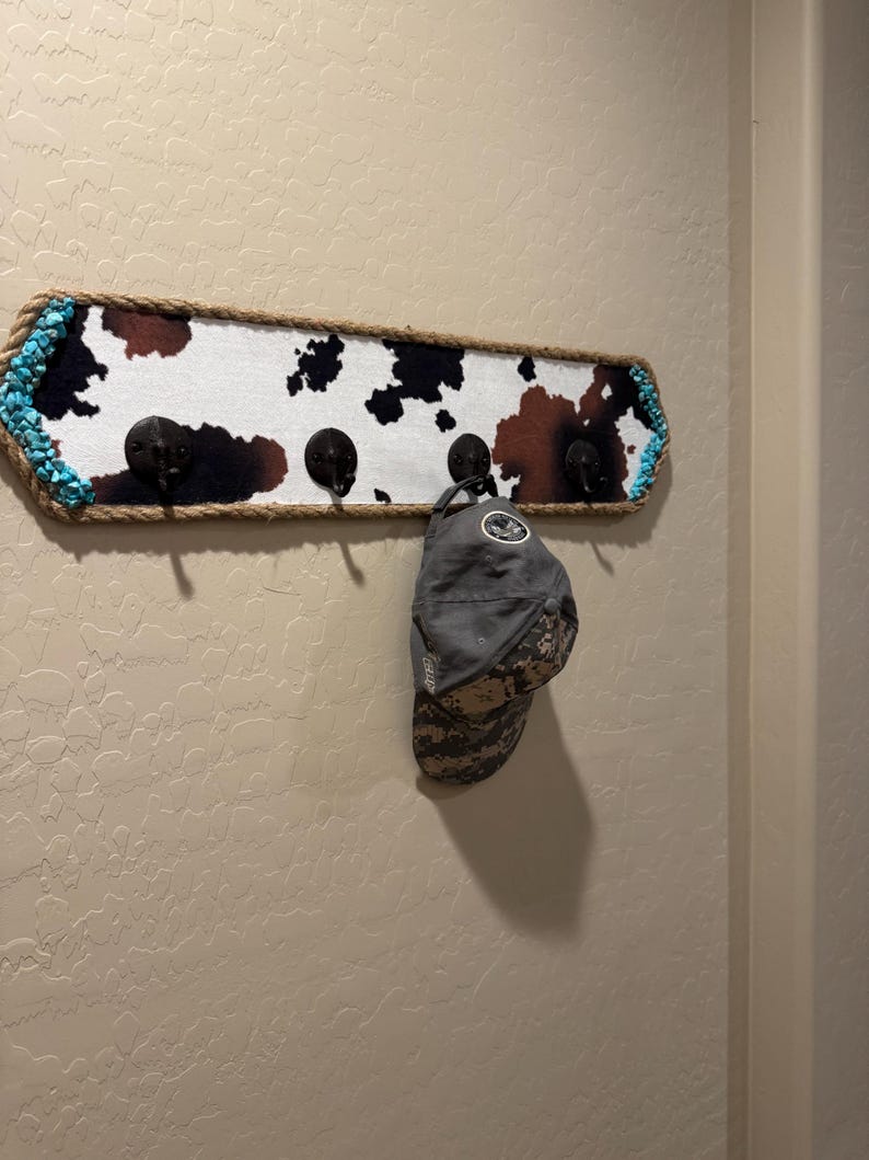 May include: A rectangular wall-mounted coat rack featuring a cowhide print pattern in black, brown, and white. The frame is wrapped in thick rope and accented with turquoise stones on the ends. Four black metal hooks hold a grey baseball cap.