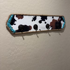 May include: A rectangular wall-mounted coat rack featuring a cowhide print pattern in black, brown, and white. The board is framed with thick tan rope and accented with bright turquoise stones on the left and right ends, including four black metal hooks.