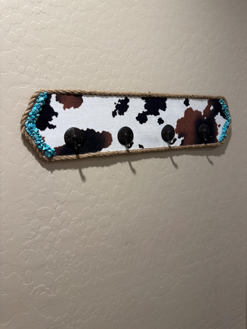 May include: A rectangular wall-mounted coat rack featuring a cowhide print pattern in black, brown, and white. The board is framed with thick tan rope and accented with bright turquoise stones on the left and right ends, including four black metal hooks.
