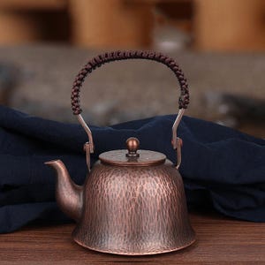 Puede incluir: Una tetera de cobre martillado con un asa trenzada. La tetera tiene un pico y una tapa con un pequeño pomo. El cobre tiene un color marrón rojizo y una superficie texturizada. El asa es de color marrón oscuro.