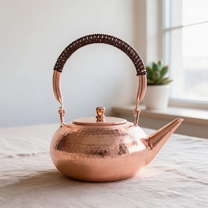 May include: A polished copper teapot with a rounded, hammered texture body, a matching lid with a small knob, and a curved spout. The teapot features a tall, arched handle wrapped in dark brown material, providing a contrast to the metallic finish.