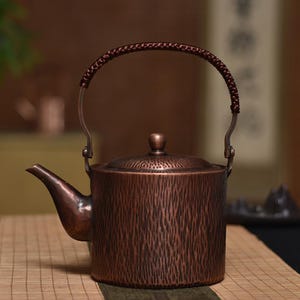 Könnte beinhalten: Eine kupferfarbene Teekanne mit strukturierter Oberfläche und geflochtenem Griff. Die Teekanne hat einen gebogenen Ausguss und einen kleinen Deckel mit einem Knopf. Ideal zum Aufbrühen von Tee.