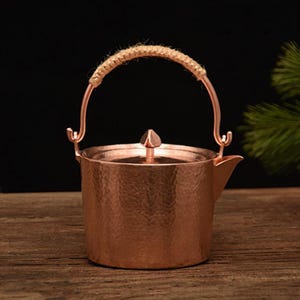 以下が含まれることがあります： ハンマー加工が施された銅色のティーポット。編みロープのグリップが付いた湾曲したハンドルと小さな注ぎ口が特徴です。蓋には装飾的なつまみが付いています。このティーポットは、お茶を淹れるように設計されています。