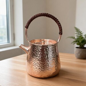 May include: A hammered copper teapot with a polished metallic finish, featuring a wide, rounded body, a short spout, a circular lid with a small knob, and a tall, arched handle wrapped in a dark brown, braided cord for a secure grip.