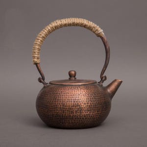 May include: A copper teapot with a hammered texture, featuring a curved handle wrapped in natural rope. The teapot has a spout and a lid with a small knob. The design is rustic and elegant.