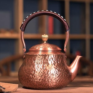 May include: A copper teapot with a hammered texture, a curved spout, and a lid topped with a gold-colored knob. The handle is brown with white stitching. The teapot is designed for brewing tea.