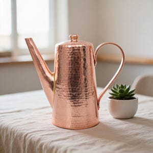 May include: A polished copper teapot featuring a distinct hammered texture across its cylindrical body, a long tapered spout, a curved handle, and a small lid knob. The metallic surface reflects light, highlighting the intricate dimpled pattern.